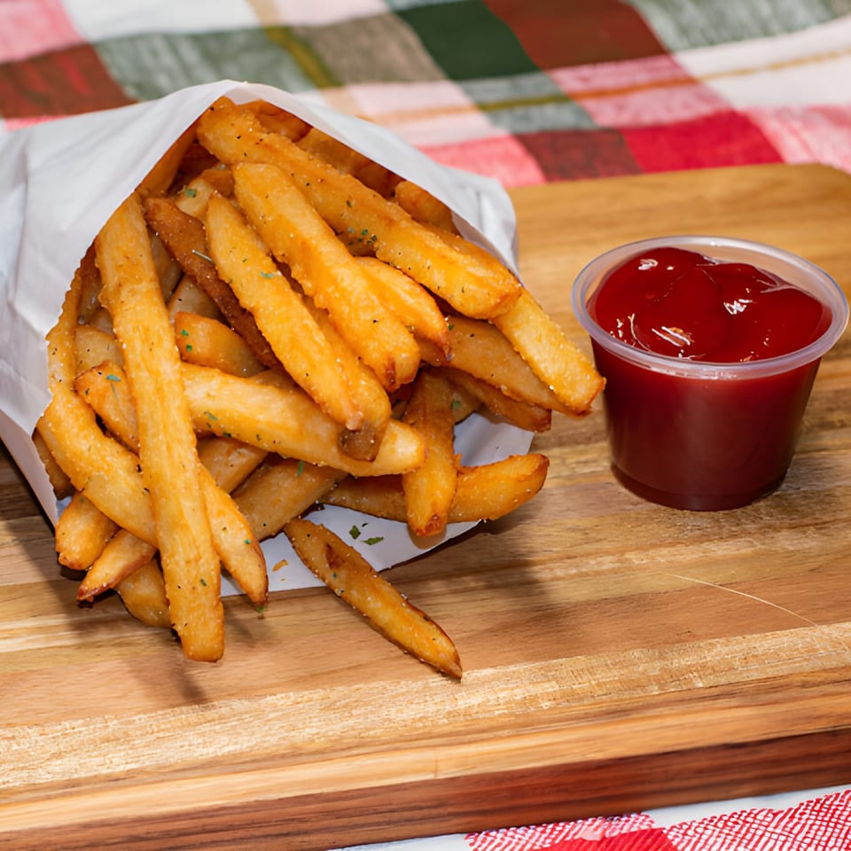 Golden french fries in a white paper bag with a side of ketchup on a wooden board.